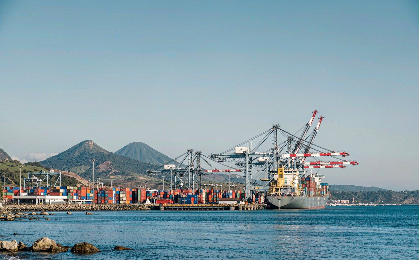 A container terminal on Guatemala's Pacific coast in clear morning light, with a container ship at the quay, gantry cranes, rows of stacked containers, calm water in the foreground and green volcanic hills in the distance
