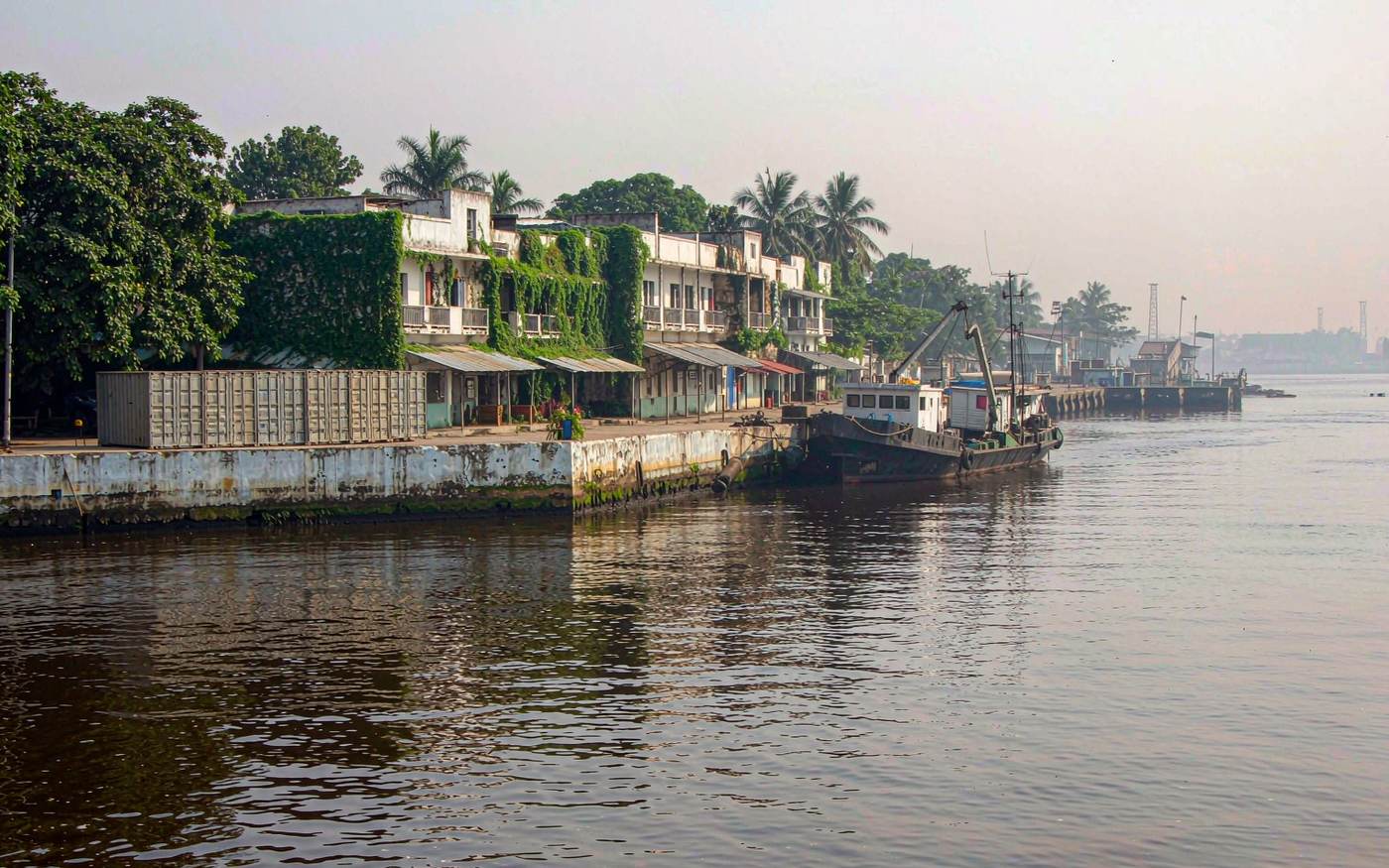 Small tidal West African river port with a simple concrete quay, a few stacked containers and a small cargo vessel alongside, low weathered colonial-style waterfront buildings, palm trees, wide muddy-brown estuary water under humid light