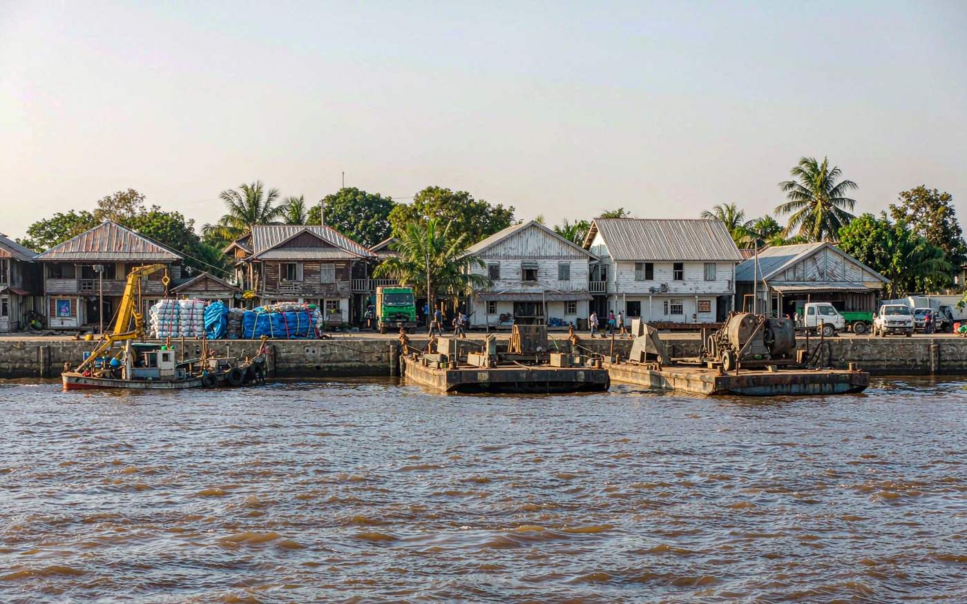 River port on a wide brown tropical river with a concrete wharf, a small cargo vessel and barges worked by a shore crane, stacked containers and colonial timber warehouses behind