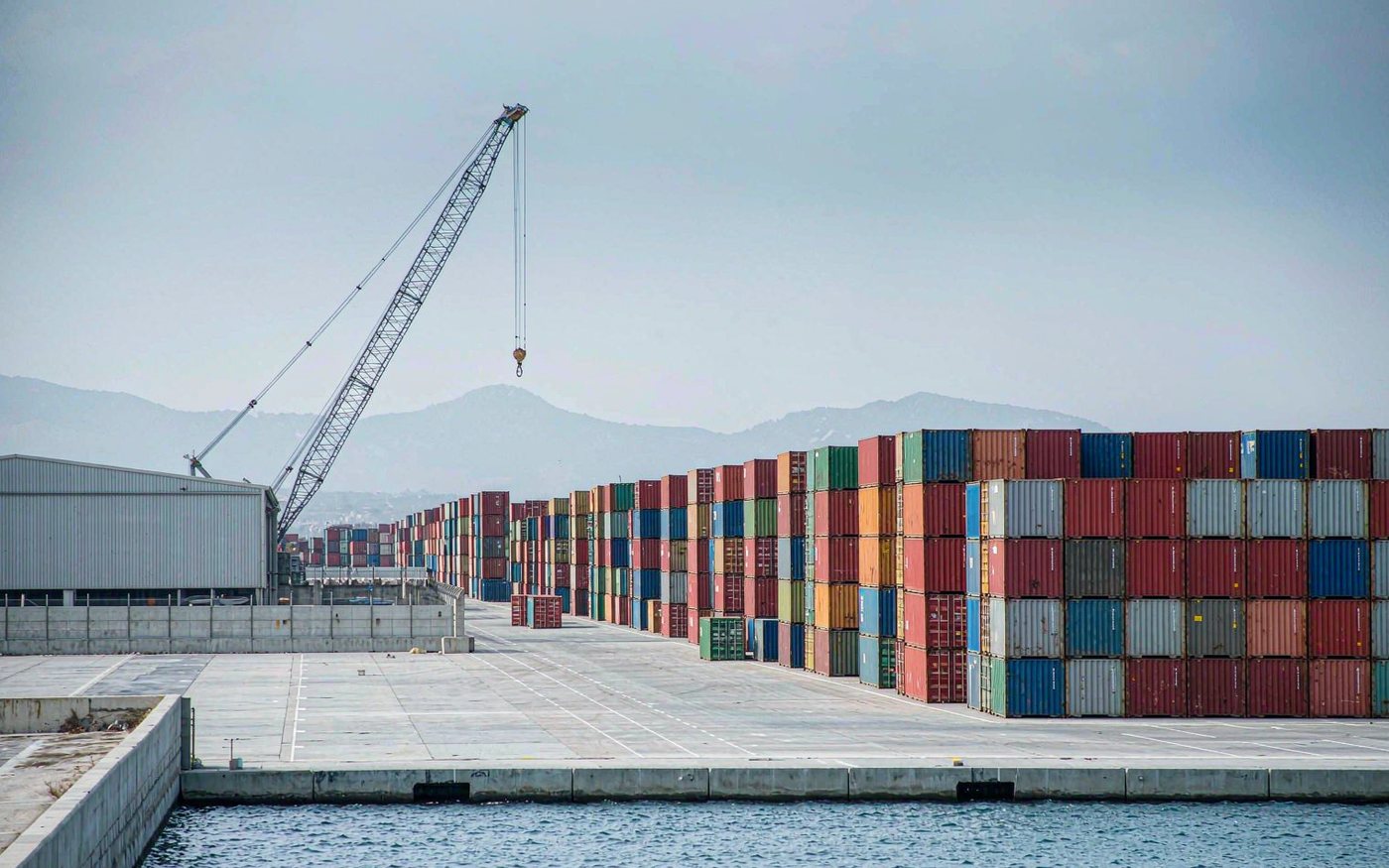 Caribbean capital container terminal with a mobile harbour crane lifting a container, stacked boxes on a paved yard and hazy mountains behind