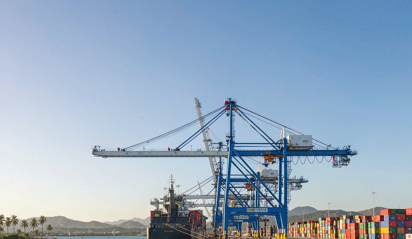 A busy Caribbean container port on Honduras's north coast in bright mid-morning light, with a container vessel berthed beneath gantry cranes, rows of stacked containers, calm blue-green water in the foreground and low green mountains on the horizon
