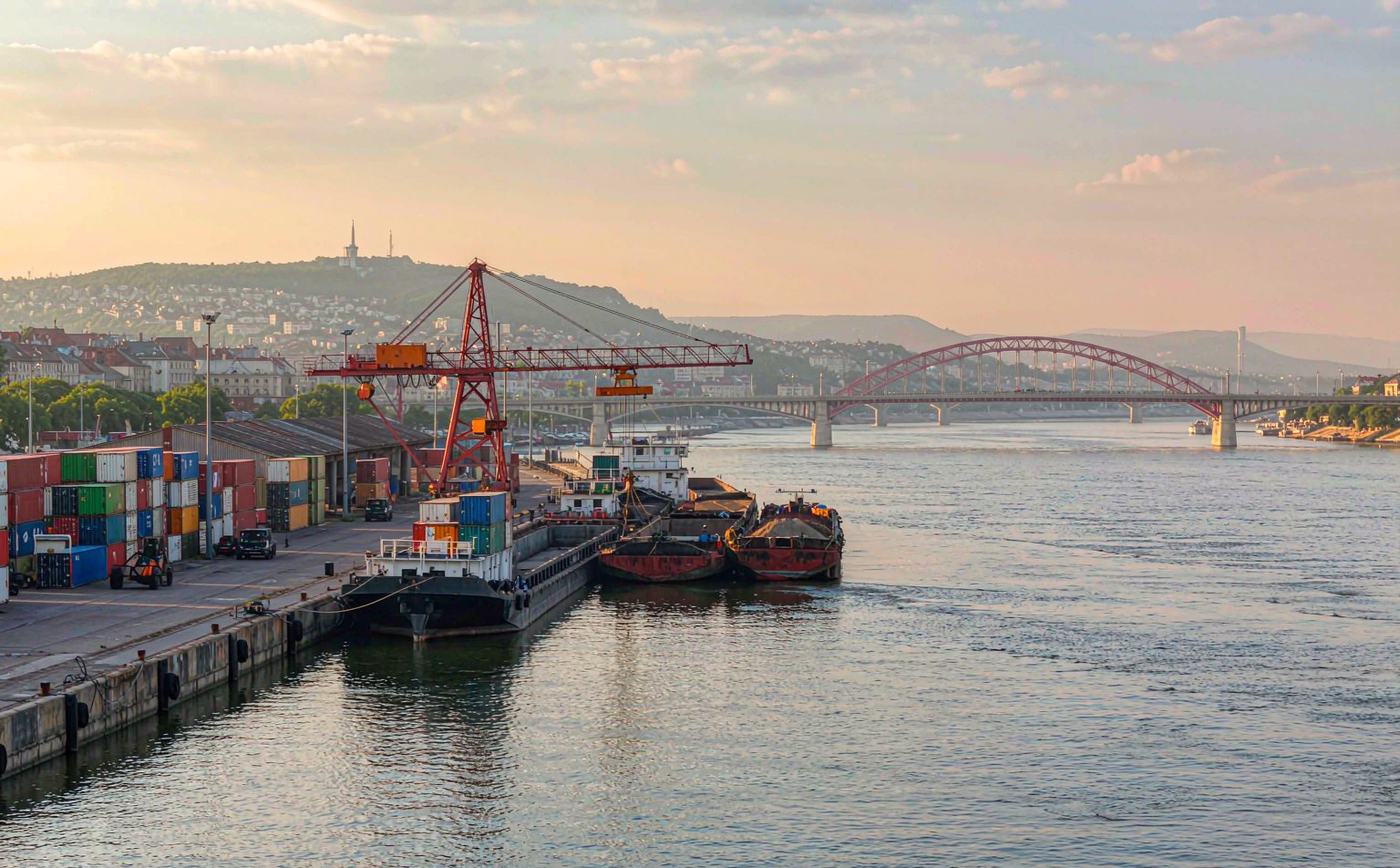 Danube river cargo port at Budapest in soft afternoon light, container and barge terminal with a gantry crane and stacked containers along the bank, low hills hazy in the distance