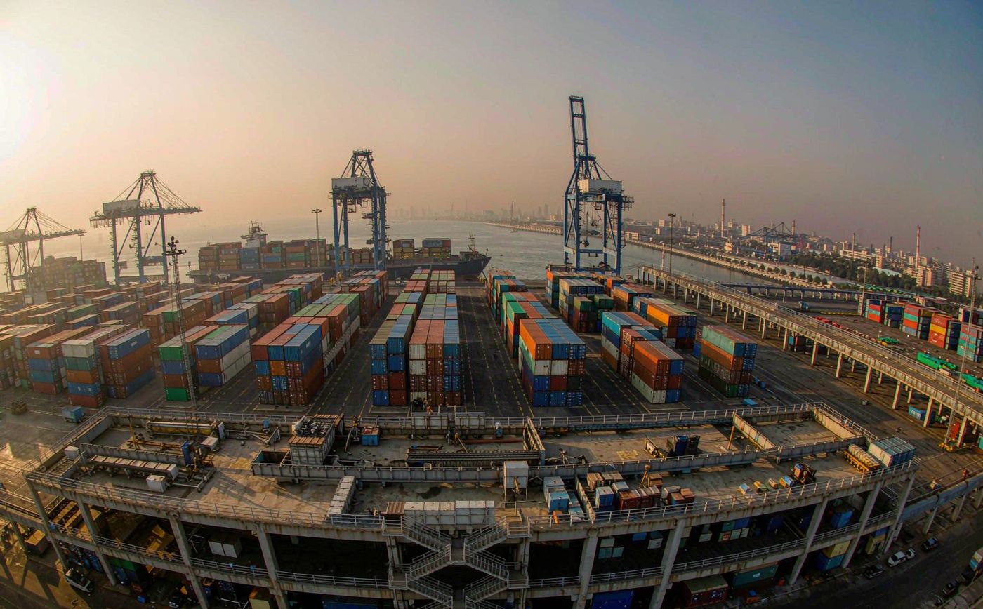 A large container terminal in India in late afternoon light, long rows of stacked shipping containers, tall gantry cranes, a container vessel berthed at the quay and hazy warm air over the port