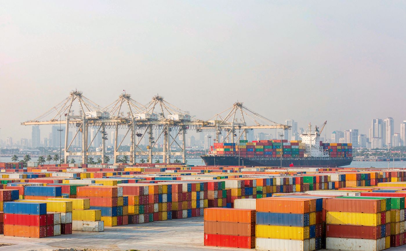 A busy Indonesian container port at mid morning, dense rows of stacked shipping containers in assorted colours, three large gantry cranes, a container vessel moored alongside, hazy tropical light and a distant high rise skyline