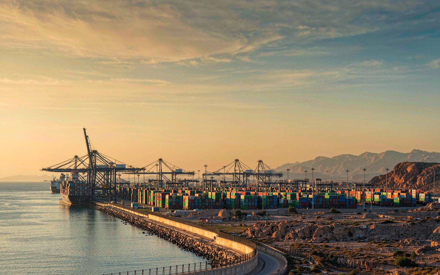 Photograph of a large container terminal on a warm southern sea coast at golden hour with stacked shipping containers in green blue and red, gantry cranes, calm harbour water and arid rocky mountains behind