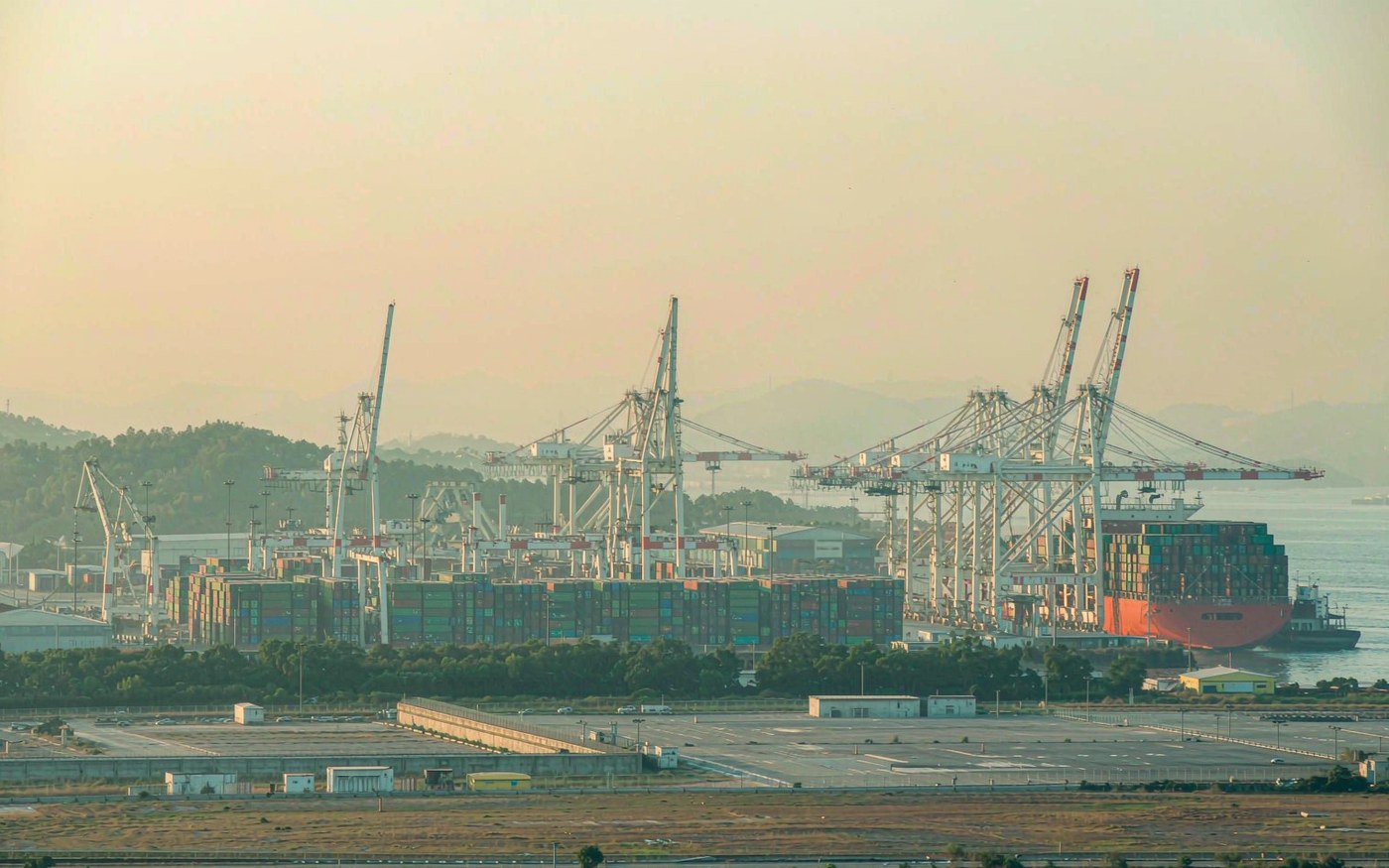 Photograph of a northern Gulf container terminal with dense container stacks and ship-to-shore cranes, a container vessel at berth, hazy warm daylight and a flat coastal landscape with low industrial buildings