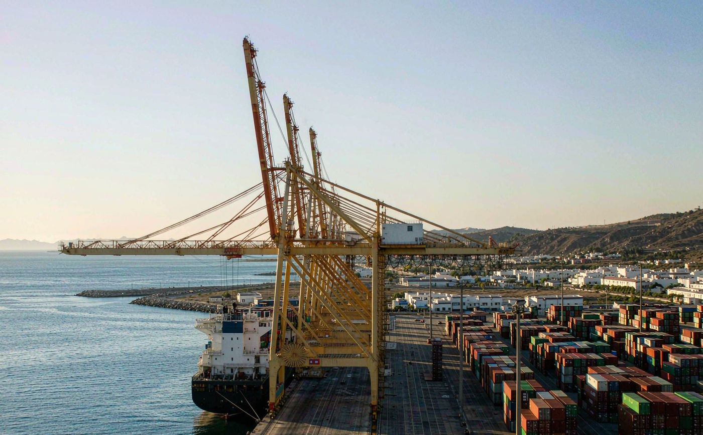 Container port on the eastern Mediterranean in bright morning light, a wide quay with tall gantry cranes, ordered blocks of stacked shipping containers, a container vessel berthed, a breakwater and open sea in front, low pale buildings and dry hills behind