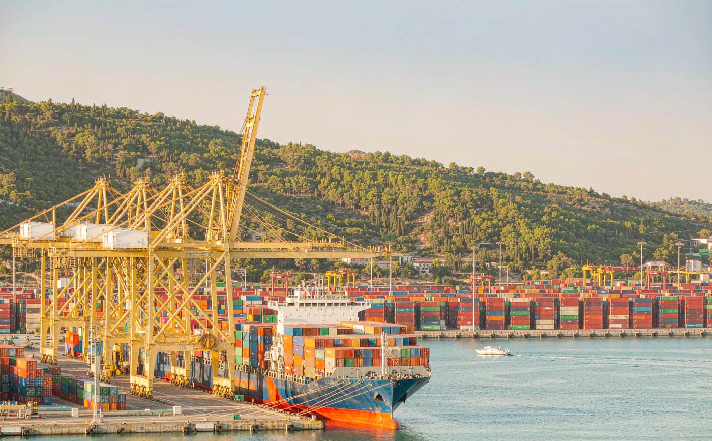 Italian Mediterranean container port with gantry cranes, dense rows of stacked containers, a vessel at the quay and hills rising behind the city