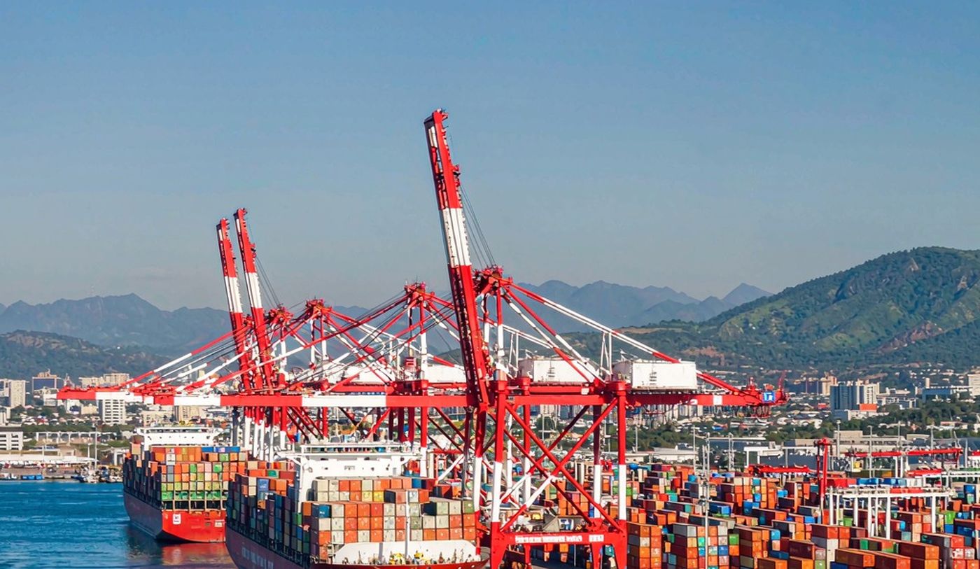 A large deep-water container transhipment terminal in the Caribbean in clear midday light, with tall gantry cranes over two container vessels, rows of stacked containers, deep-blue harbour water and hazy green mountains behind the city