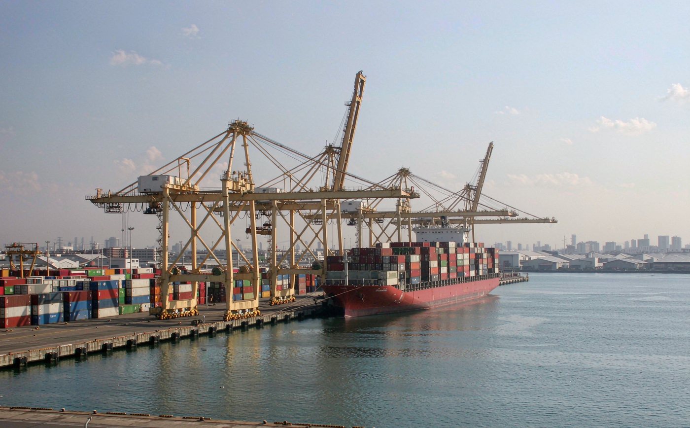 Commercial container seaport in Japan in clear daylight, a working container quay with gantry cranes and long rows of stacked shipping containers, a mid sized container vessel alongside the berth, distant low industrial buildings and a hazy city skyline, calm harbour water