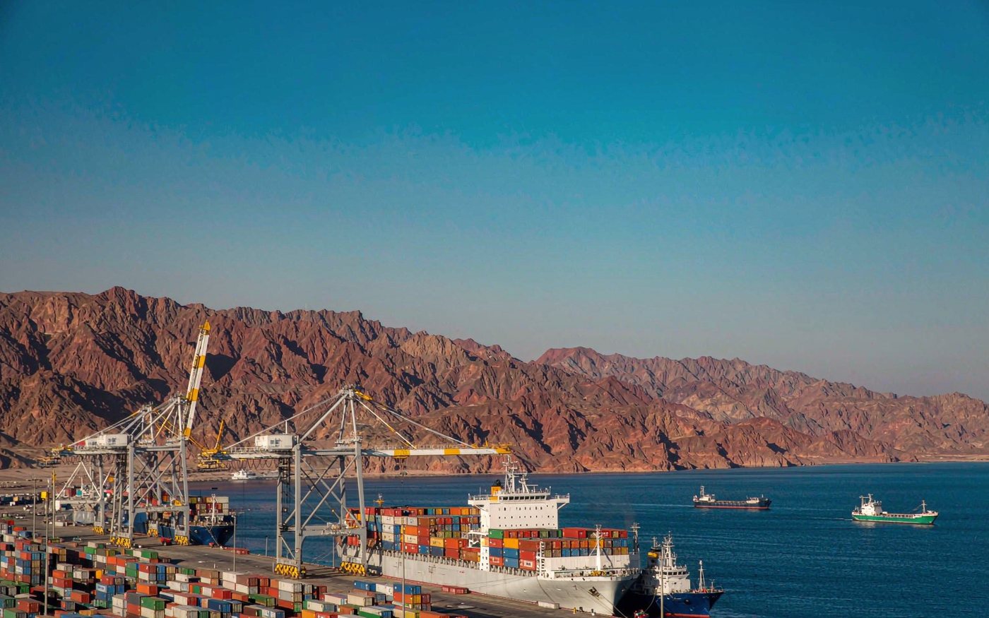 Photograph of a Red Sea container port with ship-to-shore gantry cranes and stacked containers, arid reddish mountains in the background, a container vessel berthed at the quay under clear bright daylight