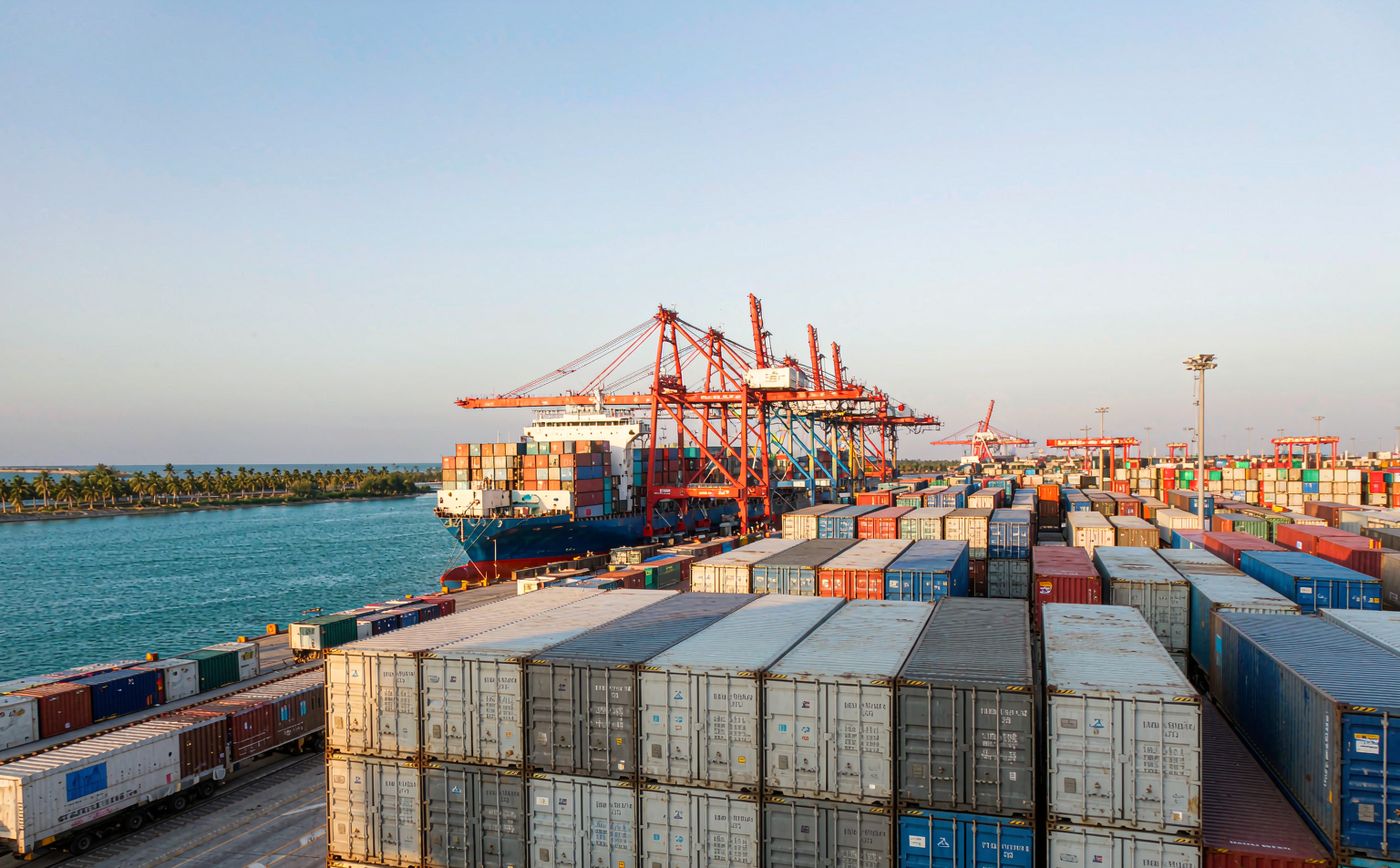 An East African Indian Ocean container port in morning light with a container vessel at the quay, portainer cranes, stacked shipping containers, a railway line beside the terminal and a tropical coastline