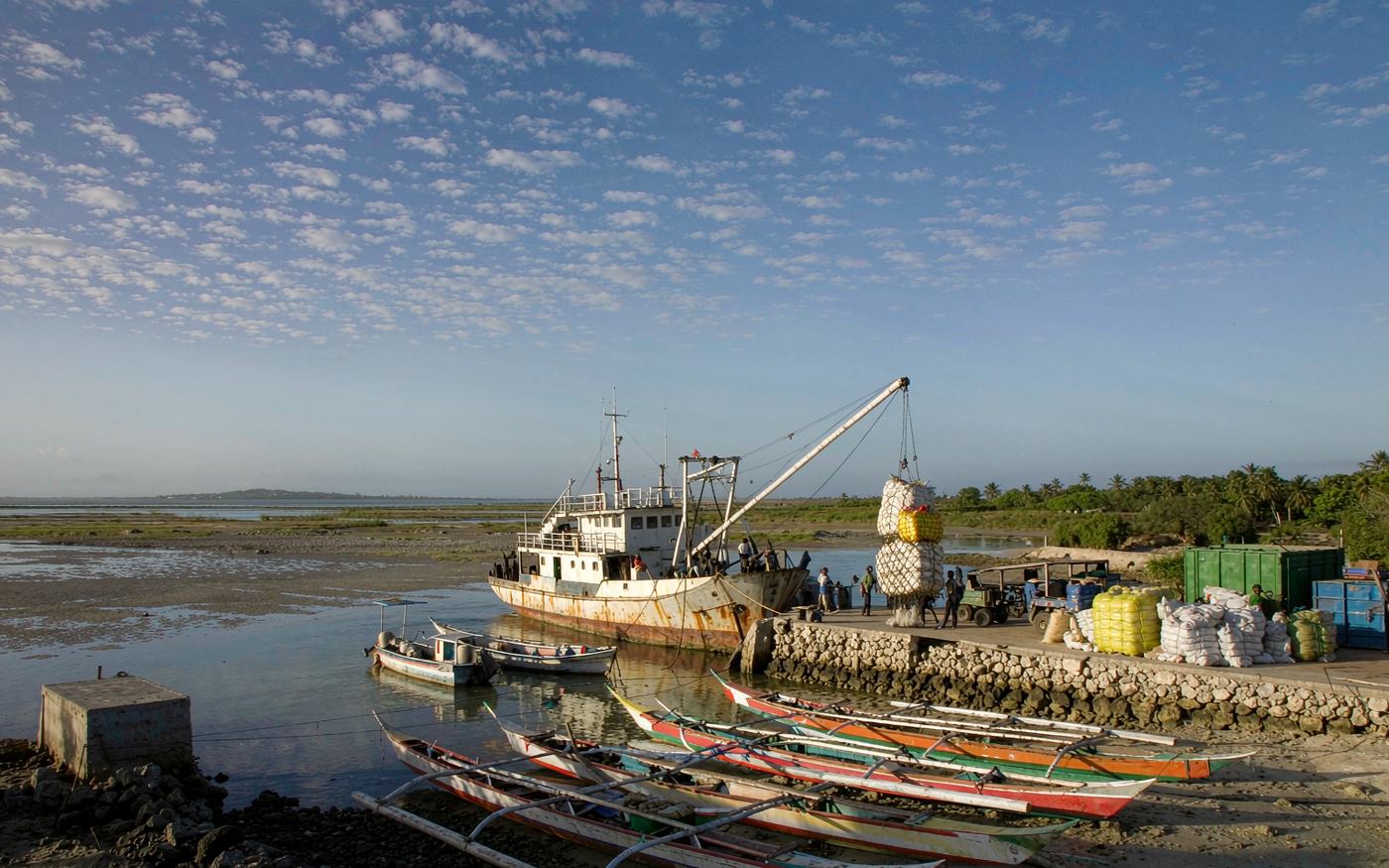 Low-lying South Pacific atoll port at low tide with a small coaster discharging bags and containers onto a coral wharf, outrigger canoes drawn up nearby and a very flat horizon