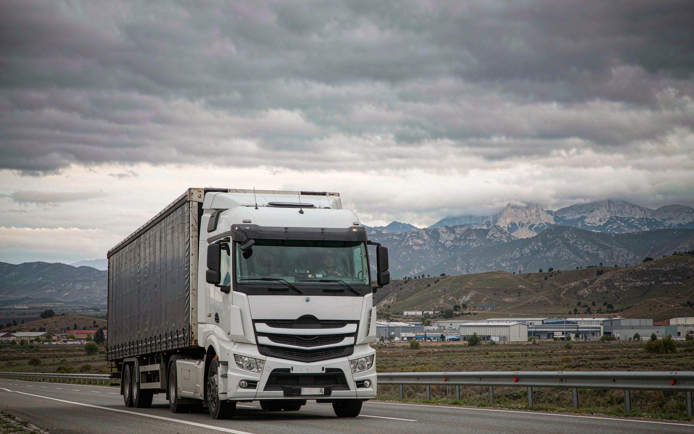 Documentary photograph of a truck carrying a shipping container on a highway through rolling green Balkan hills near Pristina, distant limestone mountains, a small industrial warehouse district, overcast central European light