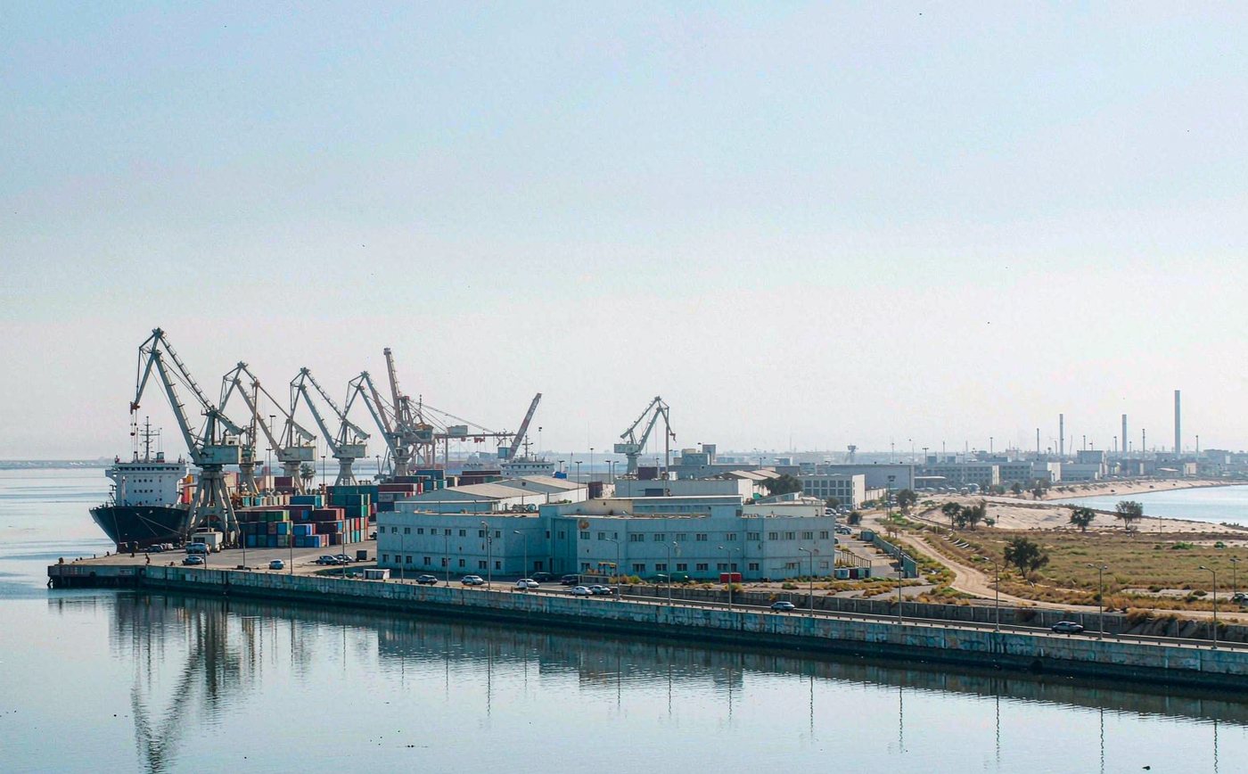 Commercial seaport on the northern Persian Gulf in clear daylight, a working container quay with gantry cranes and stacks of shipping containers, a mid sized container vessel alongside, low pale industrial buildings, calm water and a flat sandy coastline