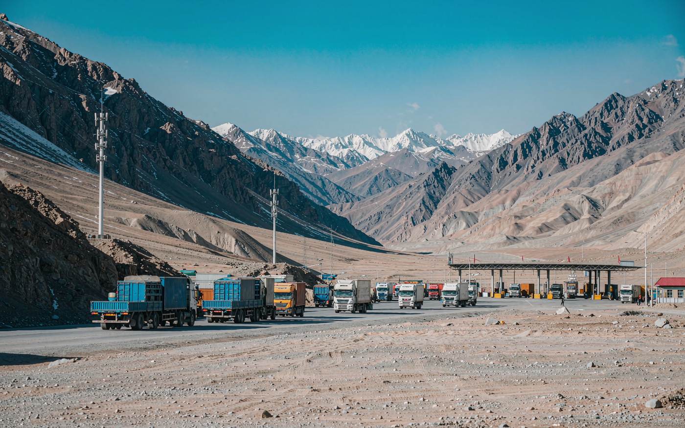 Photograph of a high mountain border crossing in Central Asia with freight trucks and containers queued at a checkpoint, snow-capped peaks and a wide valley