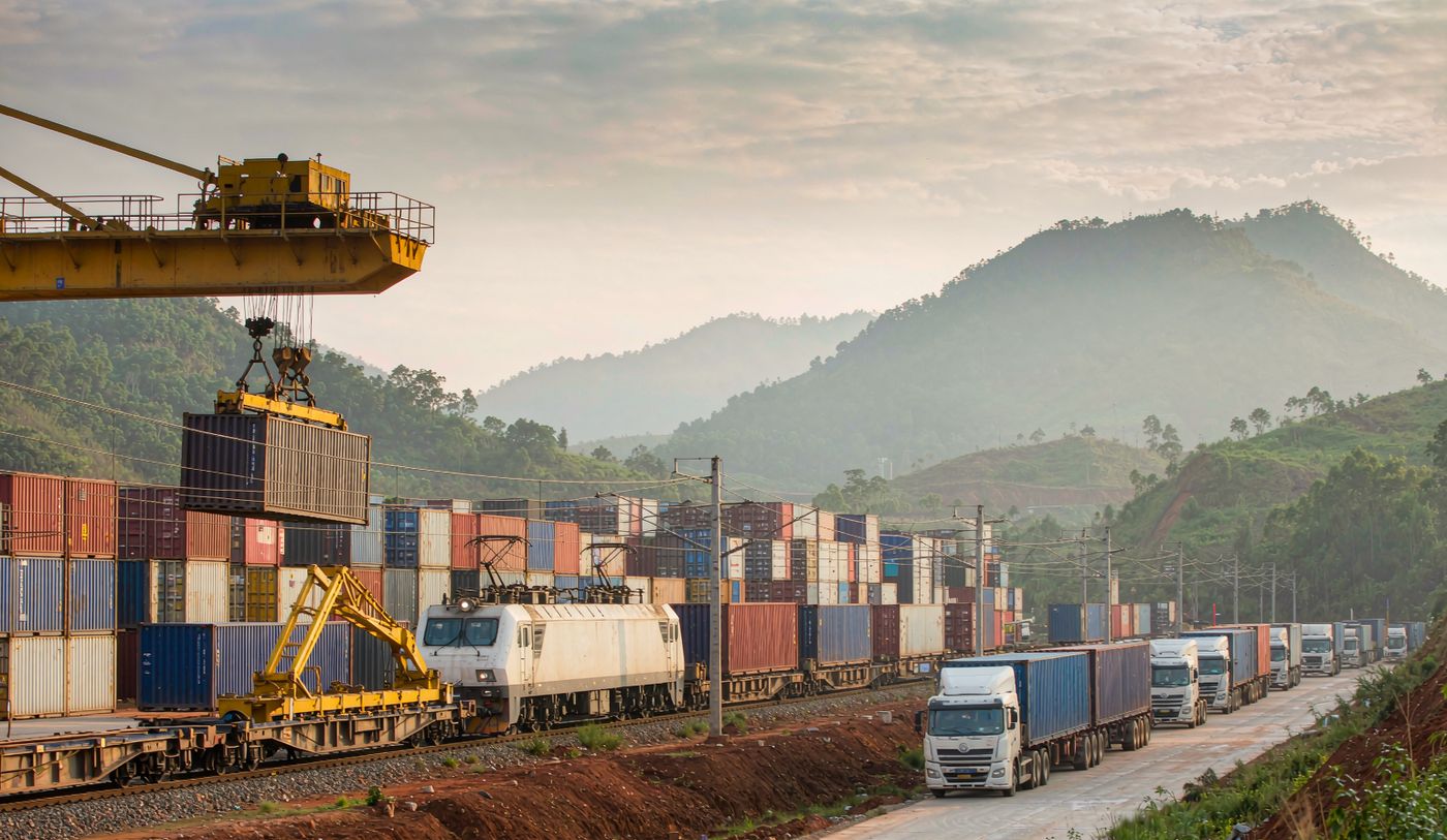 Wide photograph of the China-Laos railway at a dry port terminal near Vientiane with a container freight train on standard gauge track, gantry cranes lifting containers between wagons and stacked rows, an adjacent truck lane, red laterite earth and green mountains under humid afternoon haze