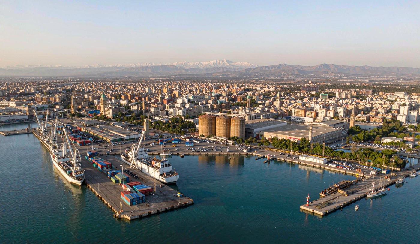 Aerial harbour view of the Port of Beirut on the eastern Mediterranean with container quay gantry cranes and stacked containers, a vessel at berth, grain silos and warehouses, blue water in the basin, low-rise city skyline and distant snow-capped mountains
