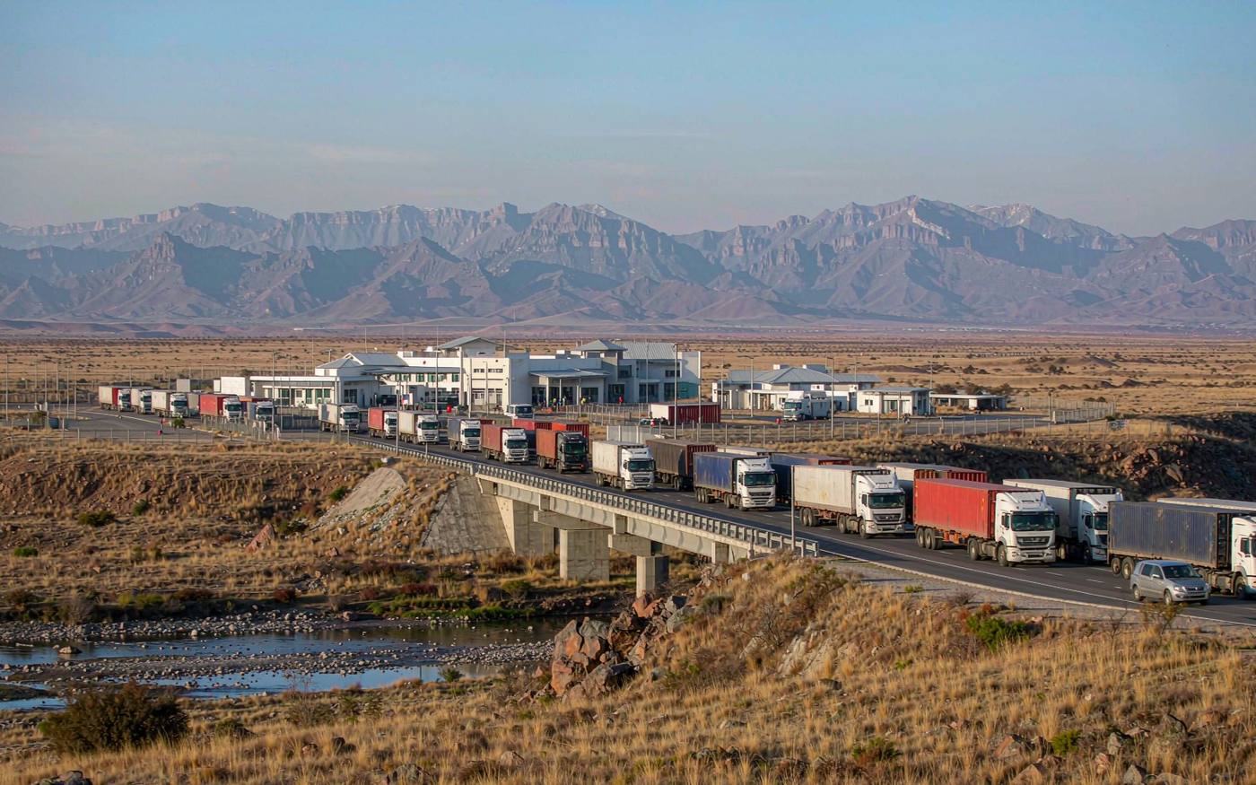 Highland border crossing in southern Africa with a wide bridge over a shallow river, queued container trucks and a border post complex, dramatic mountain ridgelines behind, dry golden grassland under thin clear high-altitude light