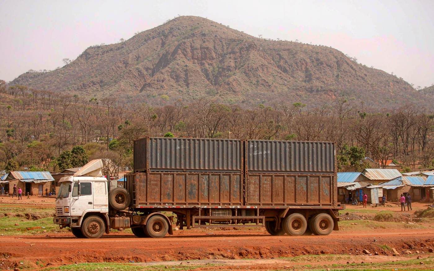 Rural southern African corridor scene with a loaded container semi-trailer on a red laterite road, miombo woodland and a distant blue mountain escarpment, a small trading settlement beside the road, warm dry-season light with dust in the air