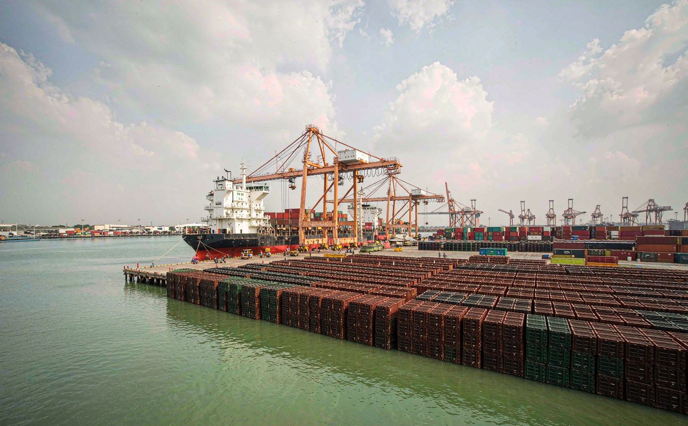 Port Klang container terminal in Malaysia in bright daylight, a working container quay with tall gantry cranes and long rows of stacked shipping containers, a container vessel berthed at the berth, tropical sky with soft clouds, calm greenish harbour water and distant low industrial buildings