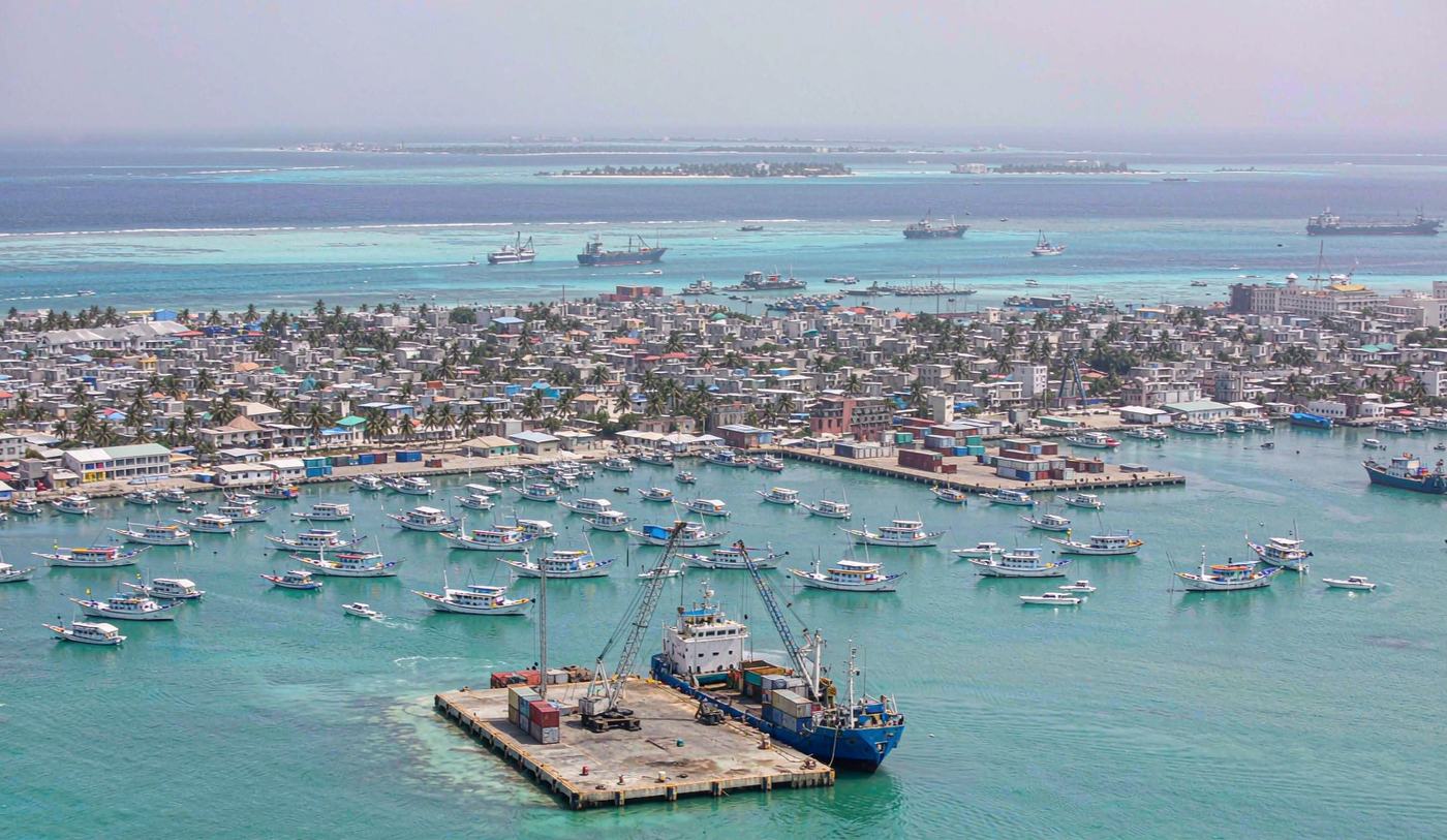 Aerial view of the commercial harbour at Male in the Maldives with a small container quay and mobile harbour crane unloading a feeder vessel, stacked containers on limited pavement, dozens of dhoni boats moored in a turquoise lagoon, dense low-rise city buildings and distant atolls under tropical midday light