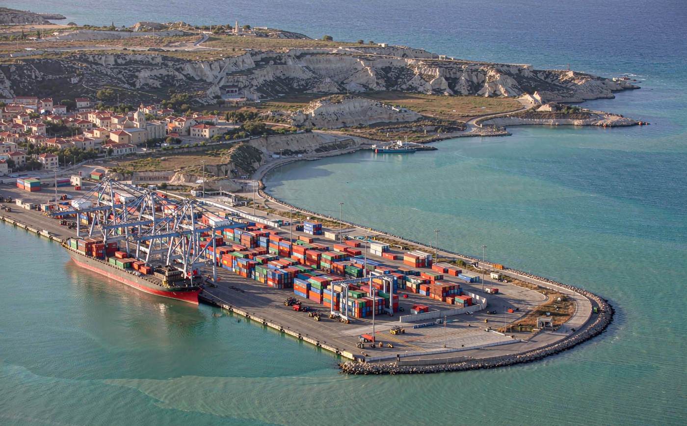 Aerial view of a Maltese container port in bright Mediterranean daylight, turquoise shallow water, a long container quay with gantry cranes, rows of stacked shipping containers, a container ship berthed and low limestone hills with a village church dome beyond