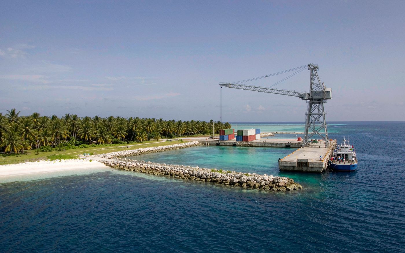 Small Pacific container terminal on a narrow coral atoll with a single ship-to-shore crane, stacked containers, a berthed feeder vessel and a turquoise lagoon behind