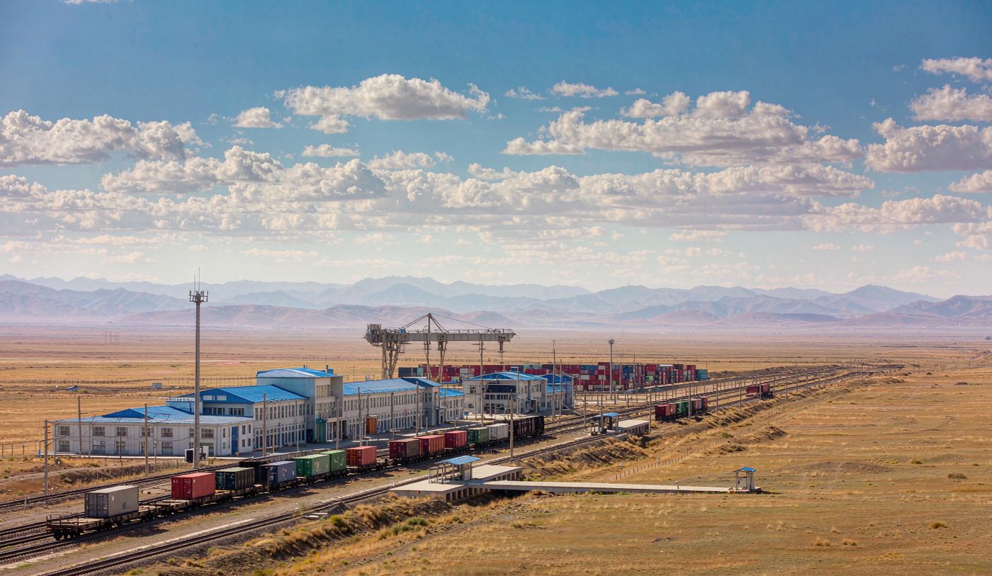 Wide photograph of a rail border crossing on the Mongolian steppe at Zamyn-Uud with broad gauge freight wagons carrying containers on parallel tracks, a gantry crane and container stacking yard, a border terminal building, flat golden grassland to a distant ridge under a vast blue sky