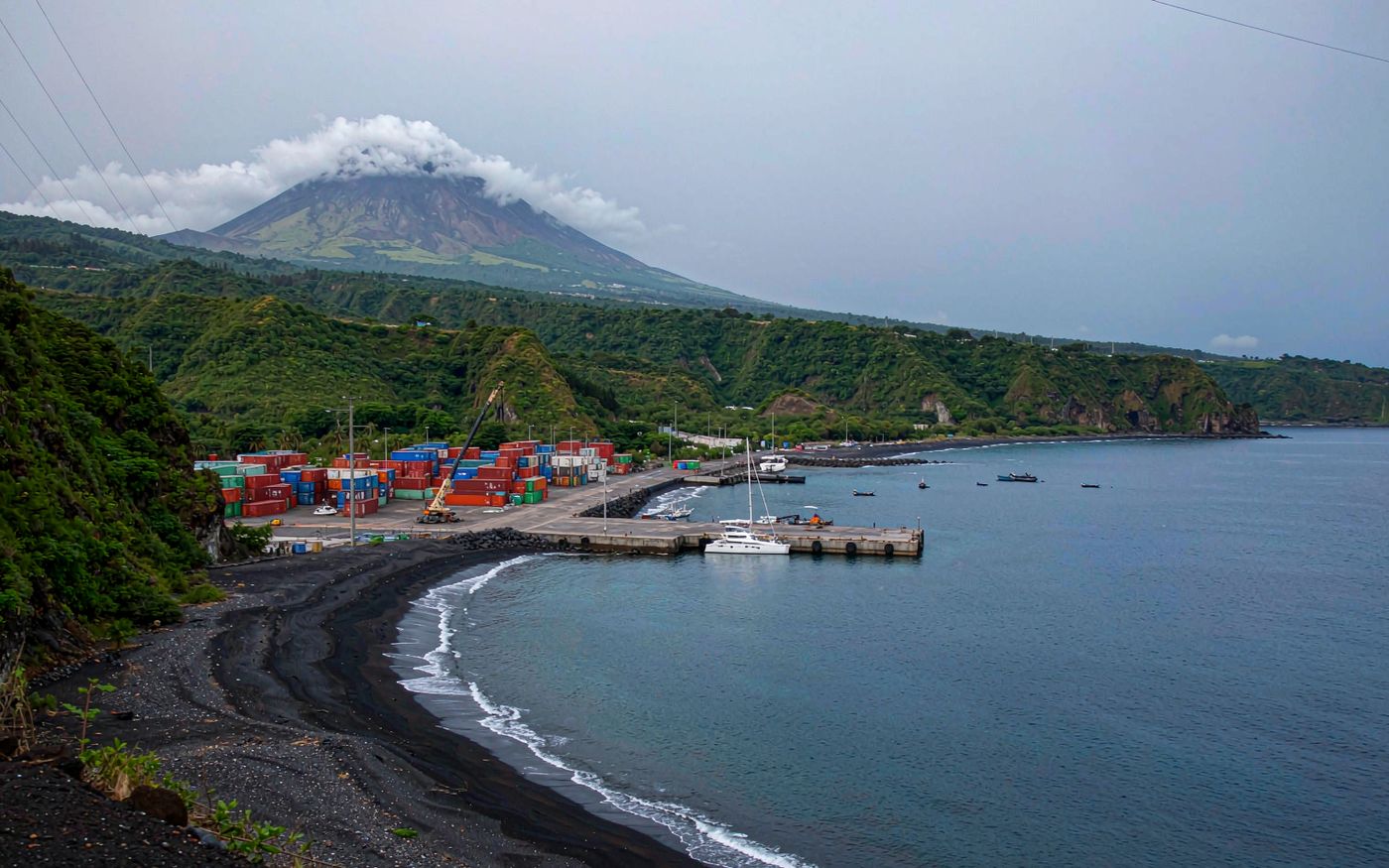 Photograph of a small volcanic Caribbean island port with a dark sand beach, a compact container yard and a single mobile crane on a small quay, lush green hills rising behind and a steep volcano peak partly covered in cloud