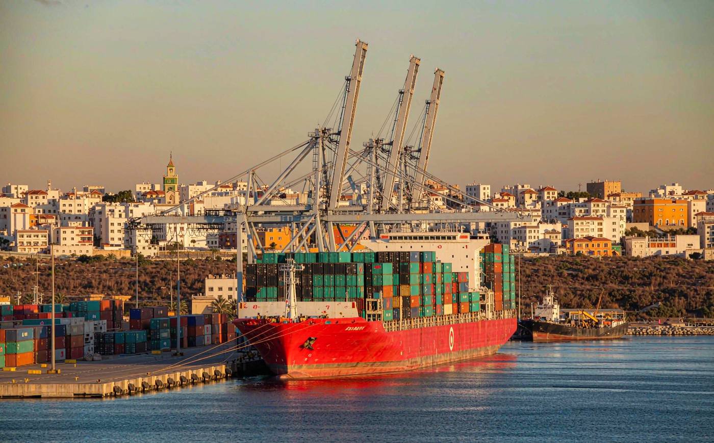 A North African Atlantic container port in late afternoon light with a container vessel at a long quay, gantry cranes, dense rows of stacked containers and a white and ochre port city on a low hill