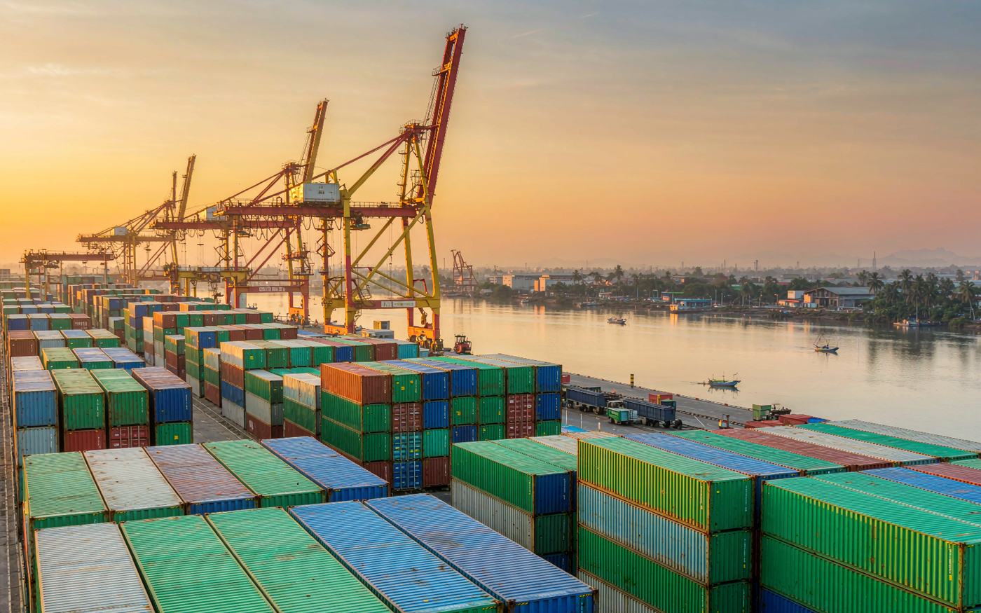 Photograph of a container terminal on a wide Southeast Asian river at golden hour with stacked shipping containers in green blue and red, gantry cranes, calm water and a distant tropical skyline