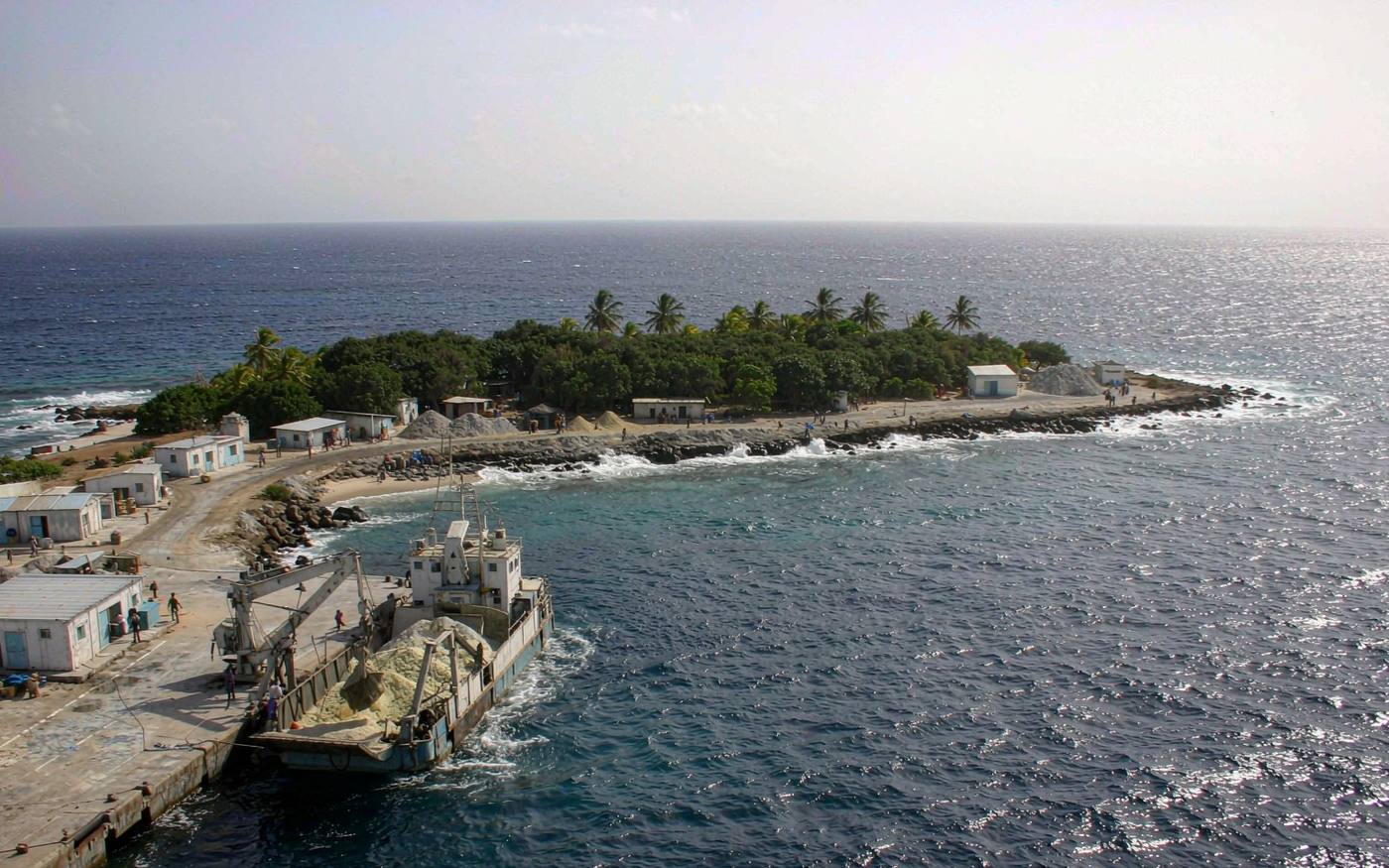 Small phosphate island harbour seen from the sea with a cargo landing craft unloading on the near shore, evergreen shrubs and palms above, deep blue open ocean beyond