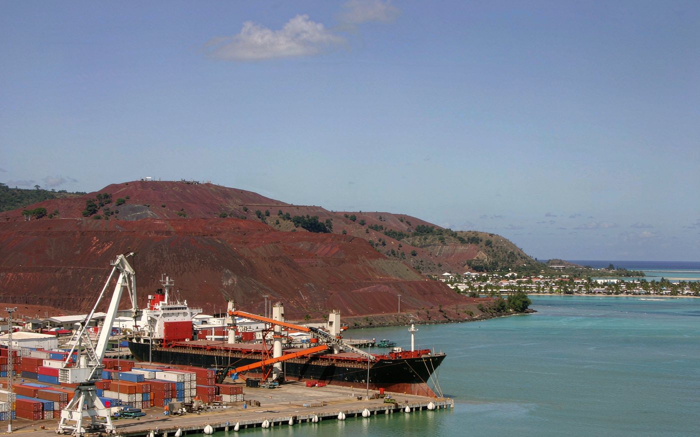 Wide photograph of a Pacific island port with a container terminal and a gantry crane, red-earthed hills rich in nickel behind, a large bulk carrier at the quay, turquoise lagoon water and a palm-lined waterfront town in the distance, bright daylight