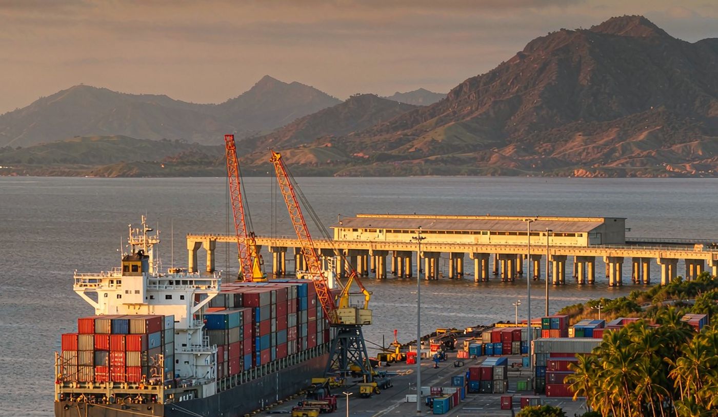 A mid-size container port on Nicaragua's Pacific coast in warm late-afternoon light, with a container vessel at the quay, stacks of containers, a long pier over calm water and green volcanic mountains on the horizon