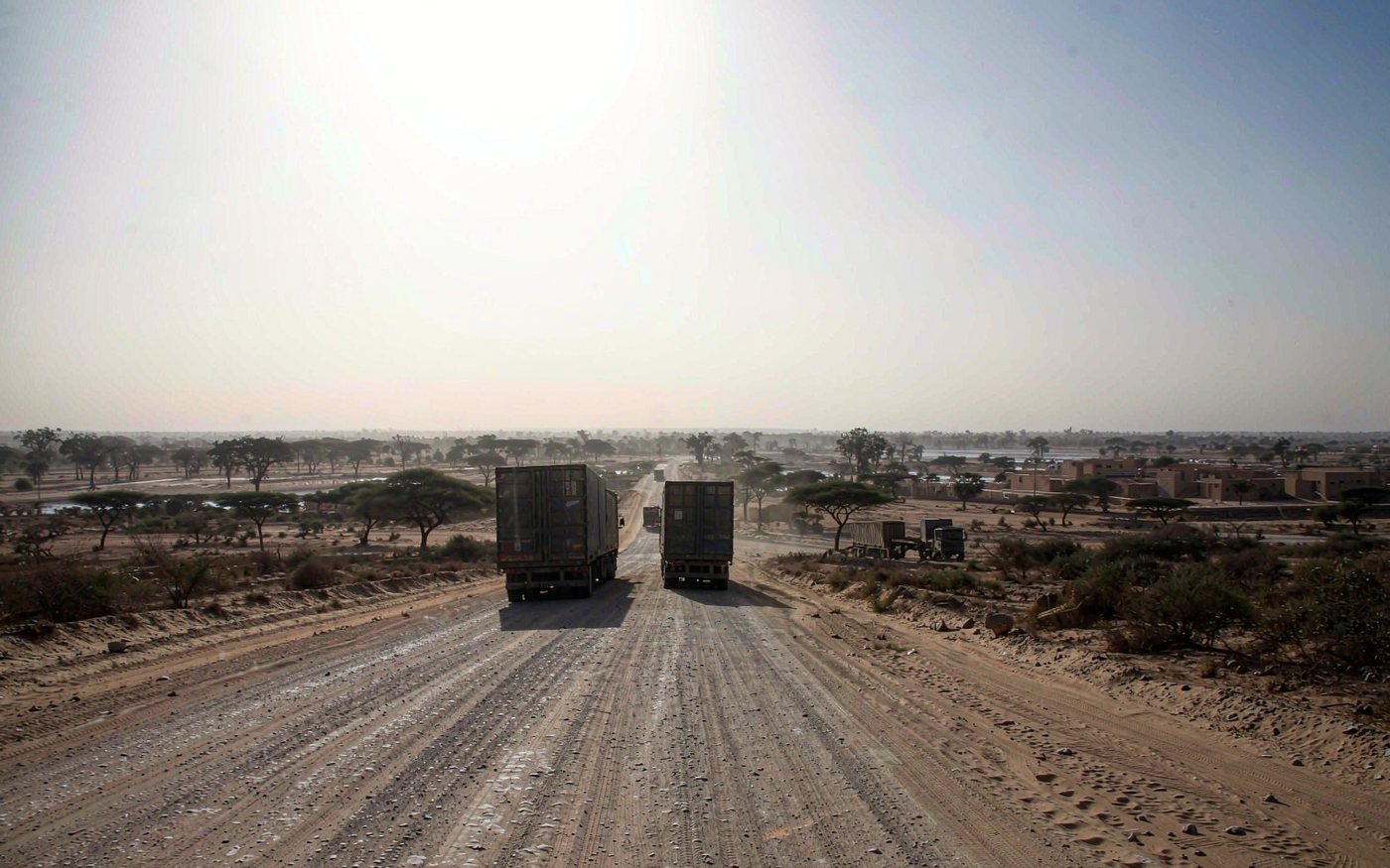 Sahel landscape near a West African capital with container trucks and trailers on a wide sandy laterite road, low acacia scrub and baobab trees, pale dusty haze on the horizon, a broad river with sandy banks beyond and low ochre buildings under bright dry light