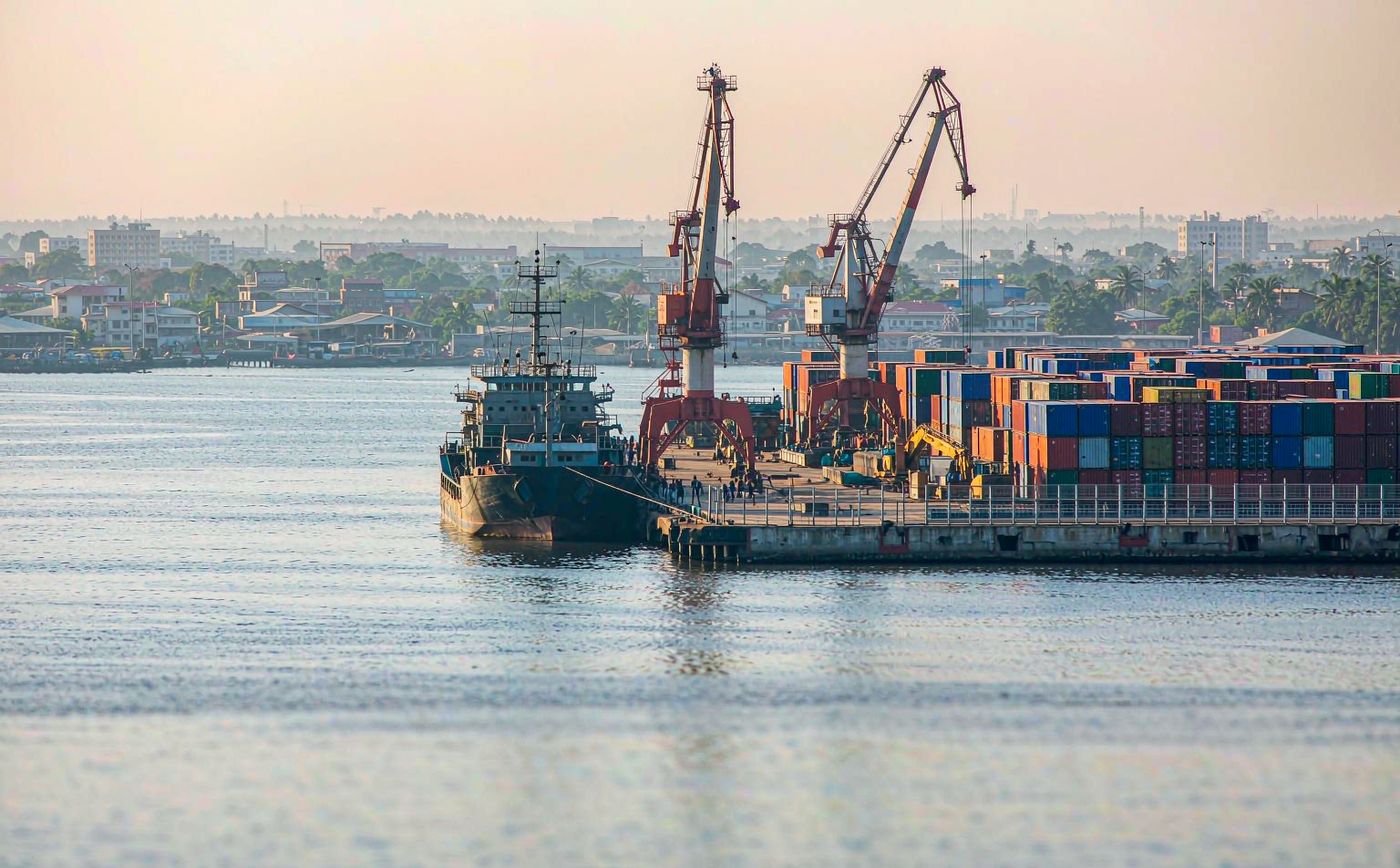 A West African container port in bright daylight with a large container vessel at the quay, gantry cranes, rows of stacked shipping containers and a tropical port skyline
