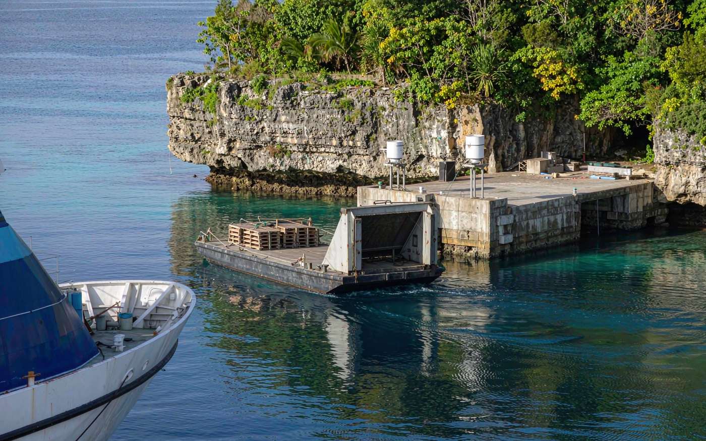 A barge carrying pallets from a ship anchored offshore toward a small wharf cut into raised coral limestone cliffs in the South Pacific