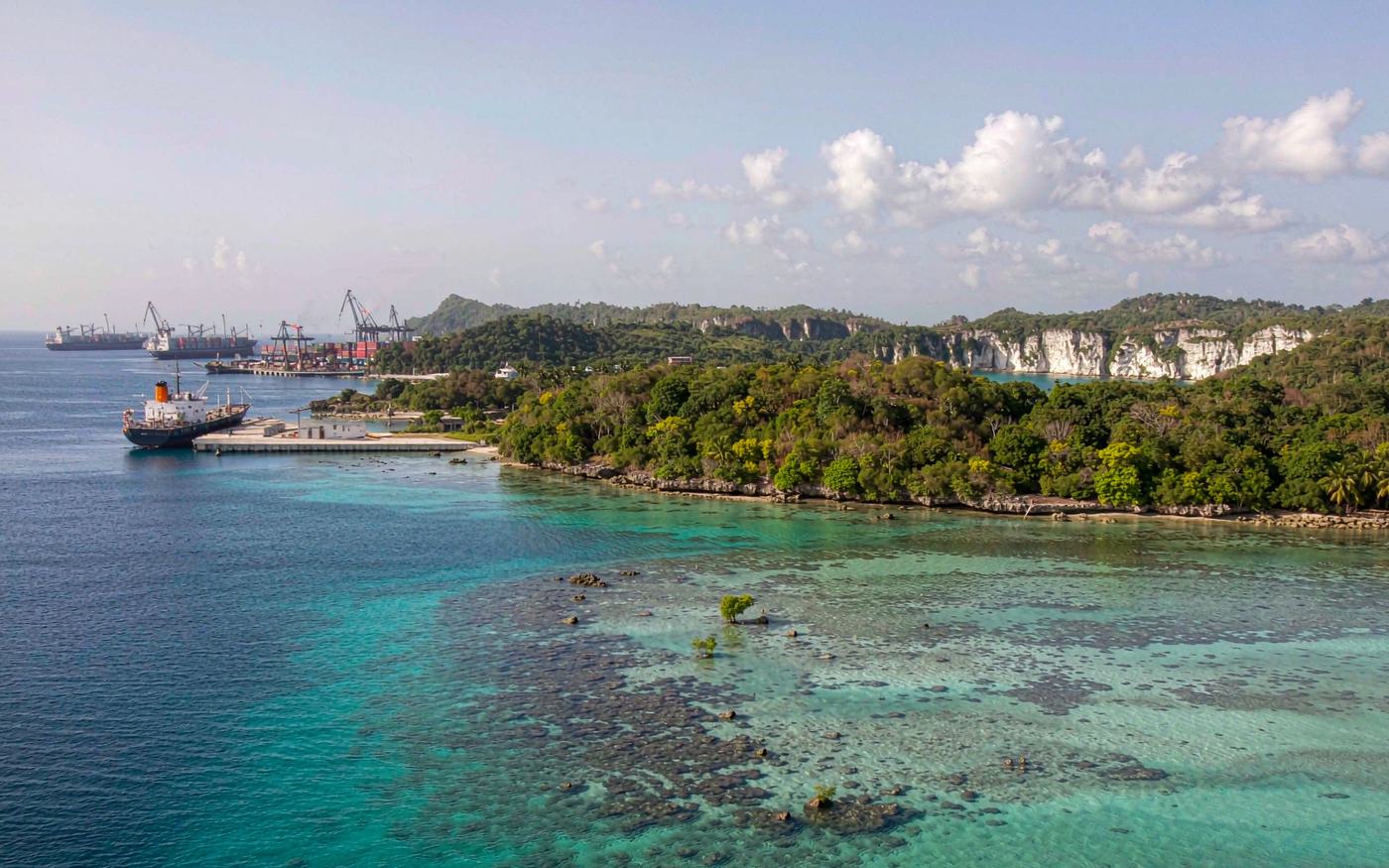 Photograph of an island port in the western Pacific with turquoise shallow water, a small cargo vessel alongside a concrete quay, container stacks, a mobile harbour crane, low green hills and white coral cliffs behind