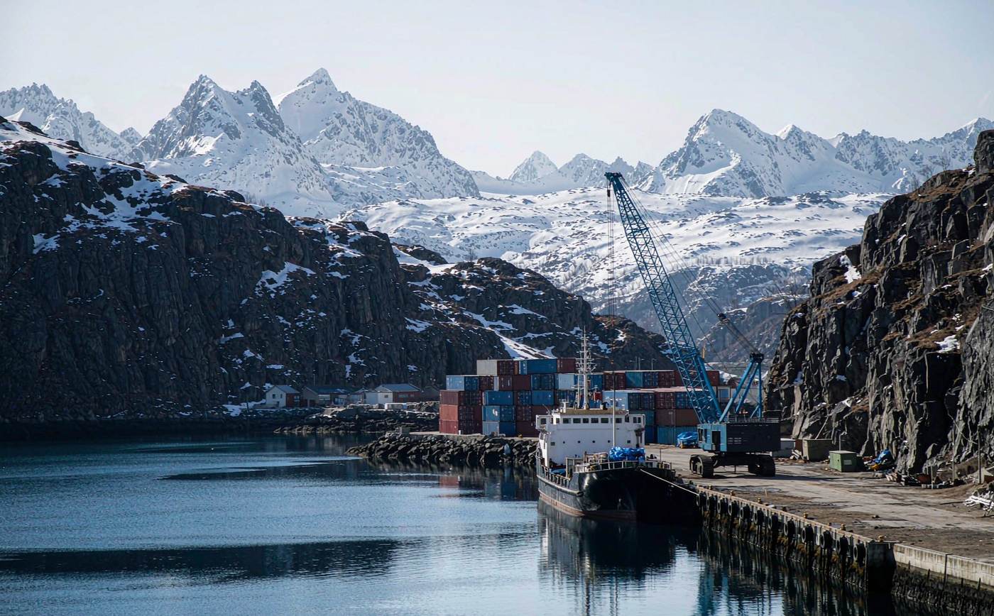 Small container and ro-ro port on a deep blue Norwegian fjord with a short-sea vessel at the quay, a mobile harbour crane, stacked containers and steep snow-patched rock slopes behind