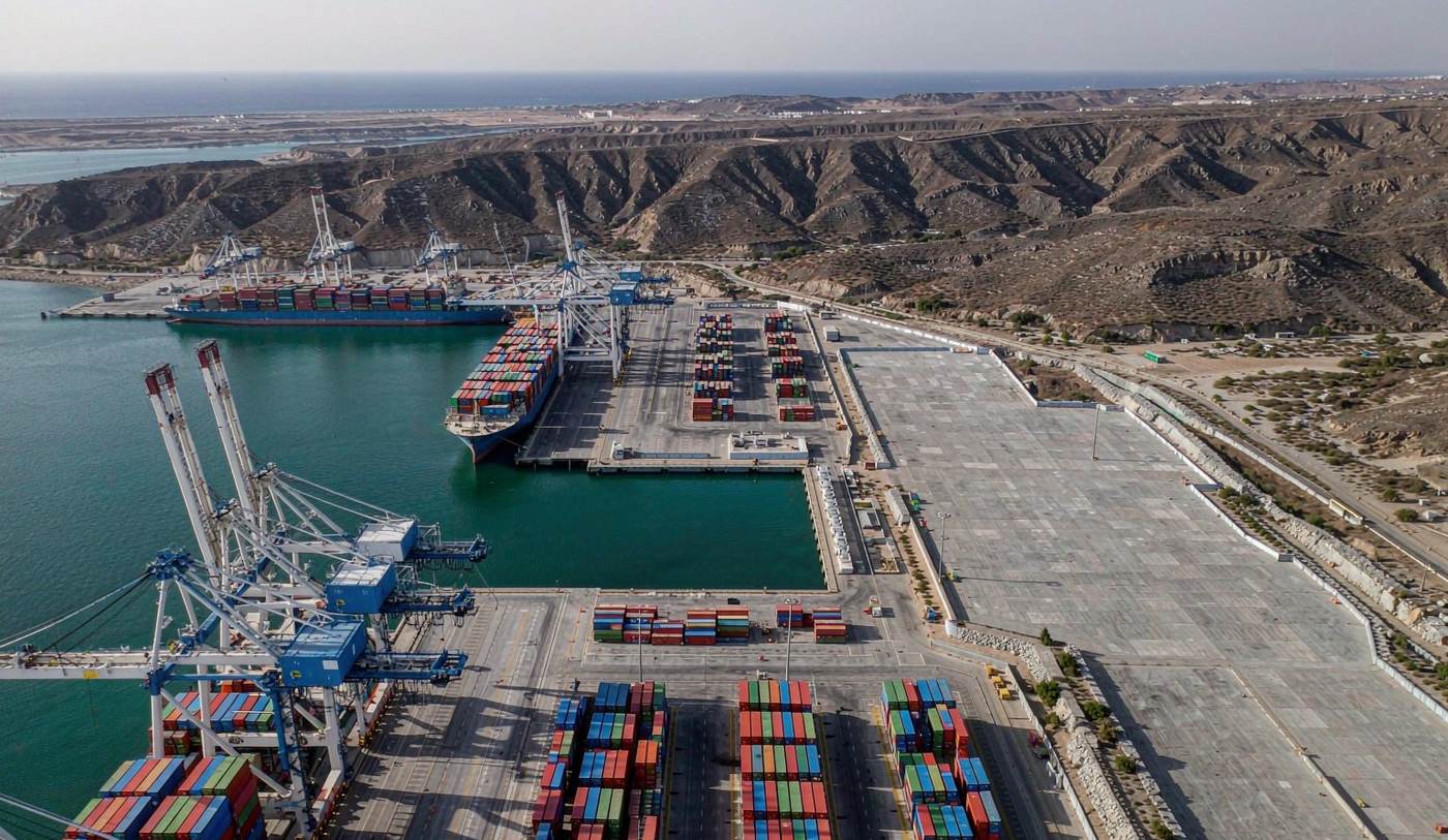 Aerial view of a modern deep-water container port on the Arabian peninsula with gantry cranes over stacks of shipping containers, a container vessel at berth, a wide paved yard, arid rocky hills and turquoise sea behind