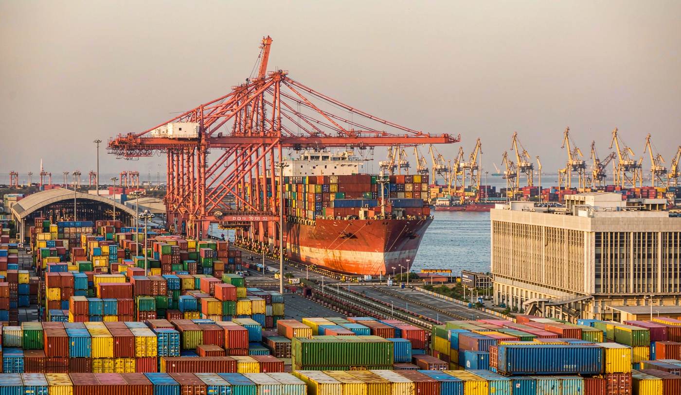 A busy South Asian container port with large gantry cranes loading a container vessel, dense stacks of shipping containers in the yard, port cranes and warehouses receding into haze under warm late afternoon light