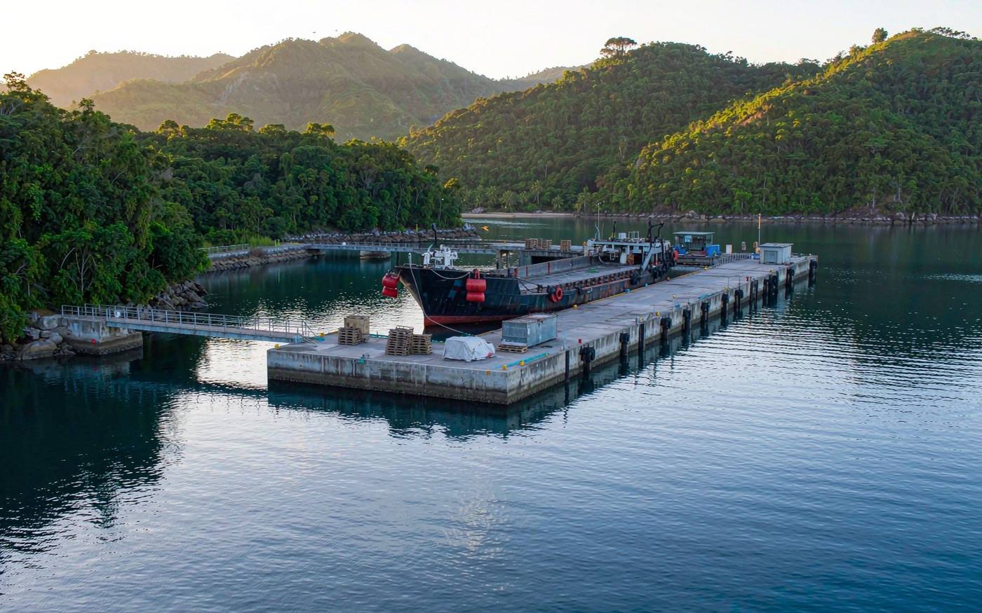 Sheltered island harbour in the western Pacific with a cargo barge alongside a short concrete quay, stacked containers and palletised general cargo, jungle-covered hills behind
