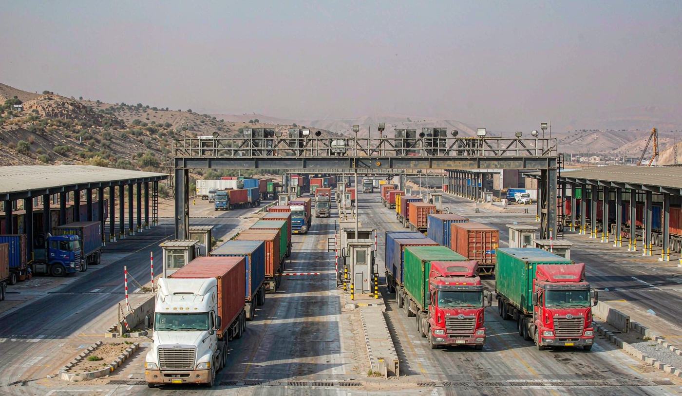 A land border cargo crossing in the Middle East with articulated trucks and shipping containers queued in lanes, an inspection gantry and warehouse sheds, arid hills and a hazy horizon behind under mid-morning light