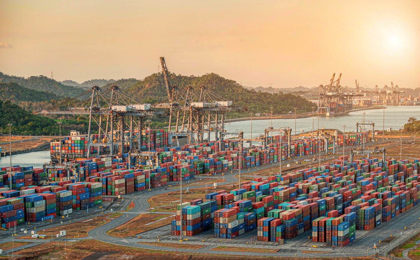 A container terminal at the Pacific entrance of the Panama Canal in early morning light, with gantry cranes loading a container ship, dense rows of stacked containers, jungle-covered hills and the calm canal channel leading towards the locks