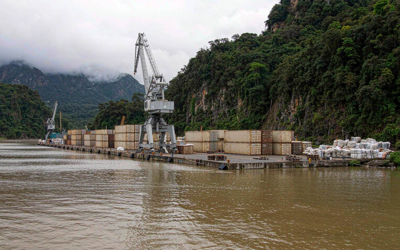 Melanesian container berth with a mobile harbour crane, stacked containers and bagged cargo on the quay, densely forested mountains behind and wide harbour water in front