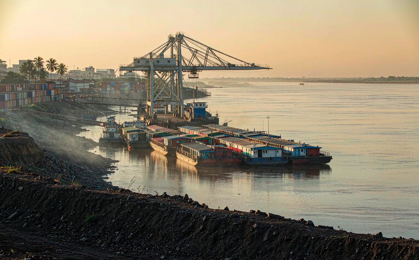A river port on the wide brown Paraguay River in early morning light, with a convoy of barges pushed by a tugboat at a small container terminal, a gantry crane, stacked containers on the bank, a low city skyline with palms and calm water to the far shore