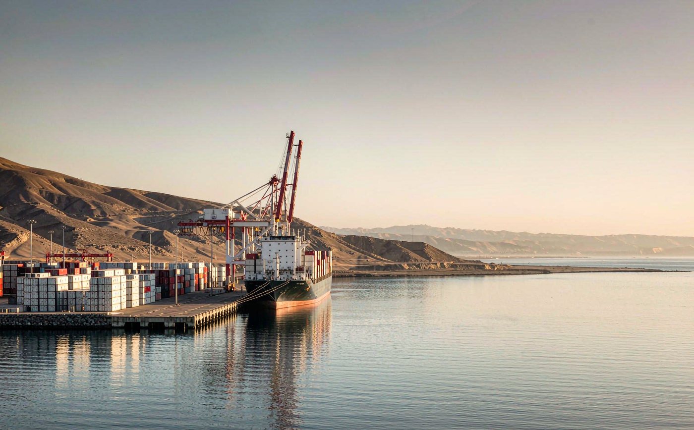 A modern deep-water container port on the arid Peruvian coast in morning light with a container vessel at a long concrete quay, tall gantry cranes, rows of stacked containers, calm water and barren coastal hills behind