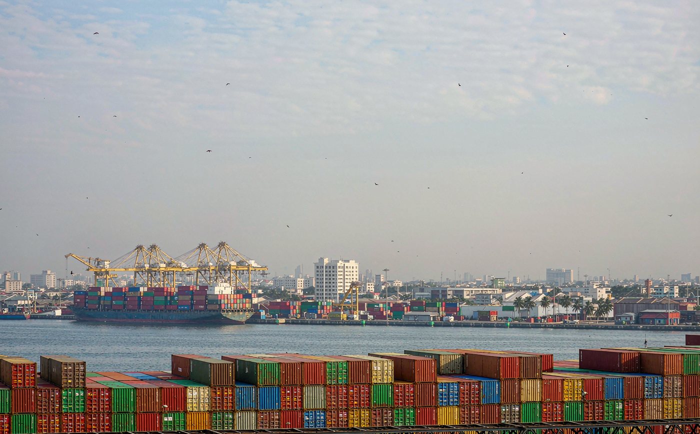 A busy container terminal on Manila Bay under bright tropical daylight, long rows of stacked shipping containers, several tall gantry cranes, a container ship berthed at the quay, hazy humid air and the Manila skyline with palm trees along the waterfront