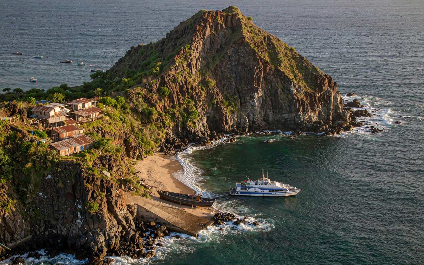 Steep volcanic island with a tiny bay, a concrete launch ramp and an open longboat, with a supply vessel standing offshore